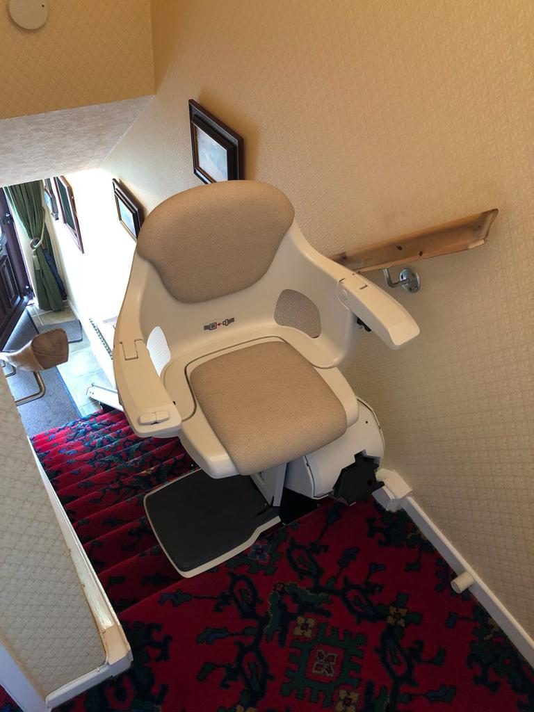 Stairlifts in Northern Ireland: Straight stairlift with a beige seat parked at the top of a red carpeted staircase
